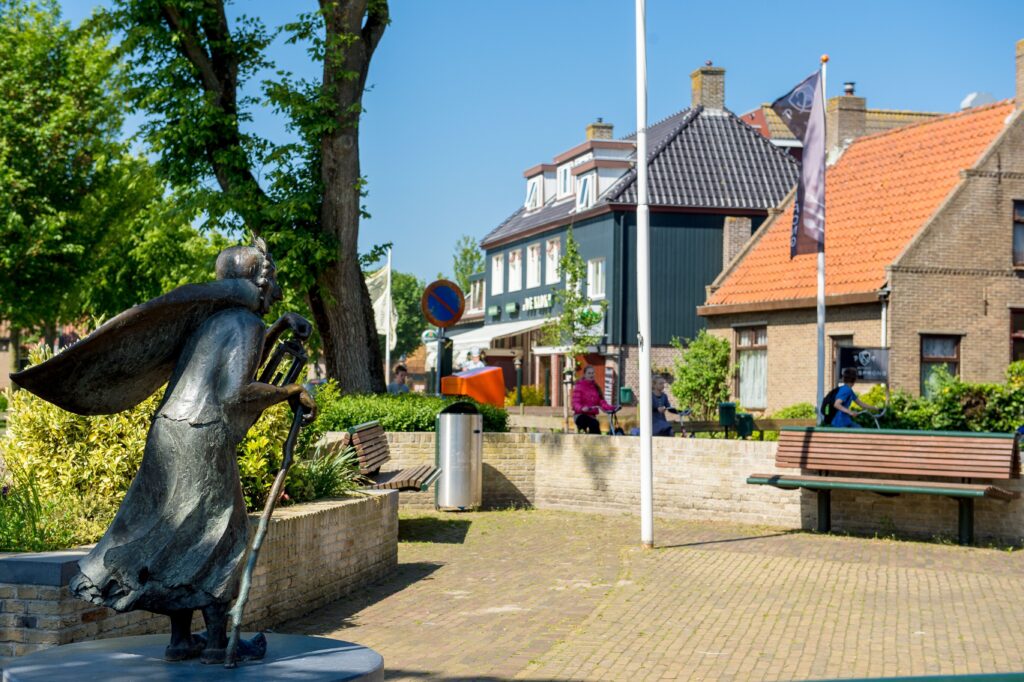 Bronzen standbeeld van man met cape op dorpsplein met traditionele Amelandse huizen en fietsverhuur op achtergrond